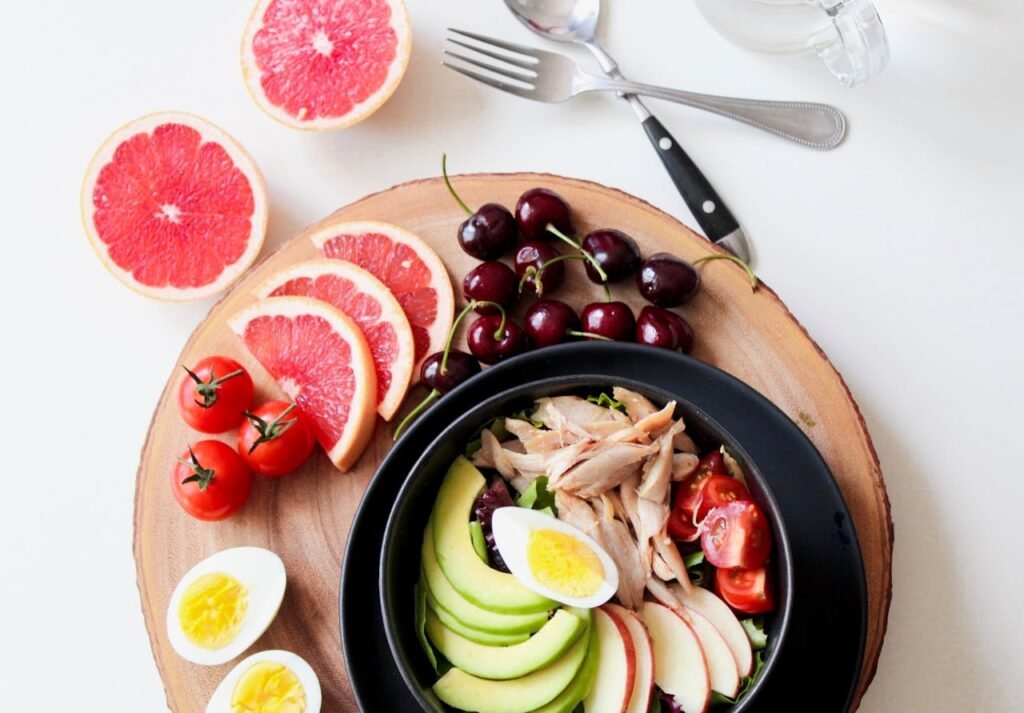 A vibrant breakfast flat lay featuring a mix of fruits, avocado, eggs, tomatoes, and sliced meats on a wooden platter.